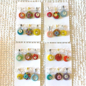 May include: A collection of handmade button earrings in various colors and designs. Each pair features a unique button, some with patterns, attached to a silver-tone earring hook. The earrings are displayed on white cards with lace trim.