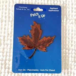 May include: A brown embroidered maple leaf patch on a blue backing. The patch is iron-on and features the text "Patch it Up". The packaging also includes the text "Appliqué" and "Iron On".
