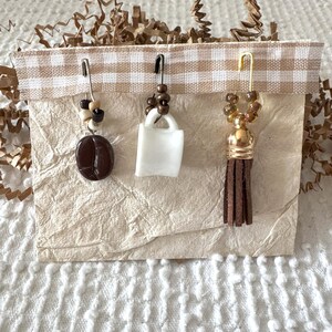 May include: Three coffee-themed charms on safety pins. One features a brown coffee bean, another a white mug, and the third a gold-toned tassel. The charms are attached to a paper card with a gingham ribbon.