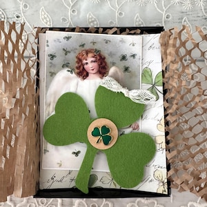 May include: A green felt shamrock with a wooden shamrock button in the center. The shamrock is in a black box with a vintage postcard of an angel with shamrocks. The box is surrounded by brown paper packing material.