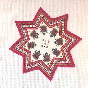 Può includere: Tessuto decorativo a forma di stella con un design vivace. Il bordo è rosa intenso, che incornicia un motivo di piccole figure in abiti colorati. Il design centrale presenta figure ripetute su sfondo bianco.