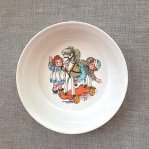 May include: A white ceramic bowl featuring a colorful illustration of Raggedy Ann and Andy riding a gray hobby horse on wheels. The graphic includes the text The Bobbs-Merrill Company Inc. and the year 1966 printed near the base of the toy horse.