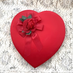 May include: A heart-shaped red box with a matching red bow and a fabric rose with green leaves. The box is sitting on a white lace background. The box is a classic gift container.