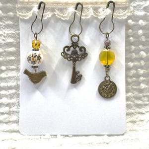 May include: Three antique-style charms on a white card. The charms include a bird, a key, and a clock face, each with decorative beads and a bronze-colored metal finish. The card is trimmed with lace.