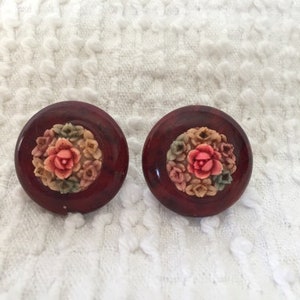 May include: A pair of red, round, floral earrings. The earrings have a raised, white floral design with pink accents.