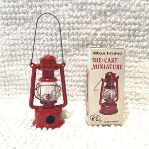 May include: A red die-cast miniature lantern with a clear glass globe and a thin metal handle, standing next to its original box labeled Antique Finished DIE-CAST MINIATURE and MADE IN CHINA, featuring a graphic of the lantern on the front.