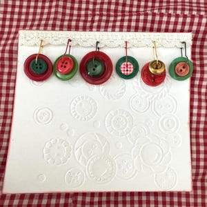 Puede incluir: Una tarjeta blanca con diseños de botones en relieve, adornada con una fila de adornos de botones coloridos. Los adornos presentan botones rojos, verdes y rosas, algunos con detalles dorados, suspendidos del borde superior. El fondo es un patrón de cuadros rojos y blancos.