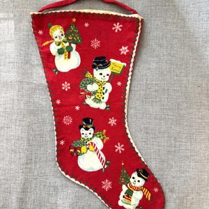 Puede incluir: Un calcetín navideño rojo con cuatro muñecos de nieve ilustrados con sombreros y bufandas, decorado con copos de nieve blancos. Un muñeco de nieve sostiene un cartel que dice Merry Xmas. Tiene bordes cosidos en verde y una asa roja.