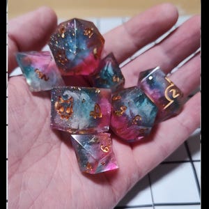 May include: A set of seven polyhedral dice with a galaxy design. The dice are clear resin with pink, blue, and gold glitter. Each die has gold numbers. The dice are held in a hand.