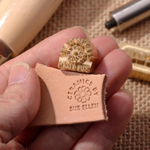 May include: A brass stamp featuring a floral design and the text CERAMICS BY SUE ELLEN, shown alongside a piece of light tan leather embossed with the same circular logo. The stamp has a metallic gold finish and a detailed engraved flower pattern.