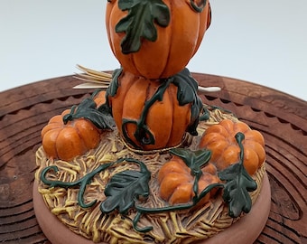 May include: A ceramic pumpkin-shaped centrepiece with three stacked pumpkins, each with green leaves and vines. The pumpkins are on a base with a straw-like texture and surrounded by smaller pumpkins.