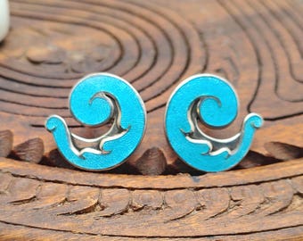 Puede incluir: Un par de pendientes de botón con un diseño de espiral en azul turquesa brillante y detalles en metal plateado. Los pendientes tienen una forma curva similar a una ola con un acabado suave y vibrante que contrasta con los bordes plateados.