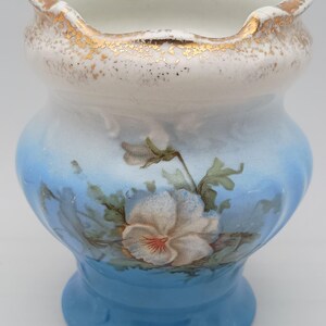 May include: A ceramic vase with a scalloped rim and a flared base. The vase is painted with a gradient of blue and white, and features a floral design with a white flower and green leaves. The rim is trimmed with gold.