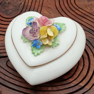 Może przedstawiać: Białe porcelanowe pudełeczko na drobiazgi w kształcie serca z wypukłym wzorem kwiatowym. Kwiaty są różowe, żółte i niebieskie.