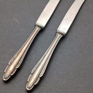 May include: Two silver butter knives with ornate handles. The knives have a polished finish and are laid out on a dark grey surface.