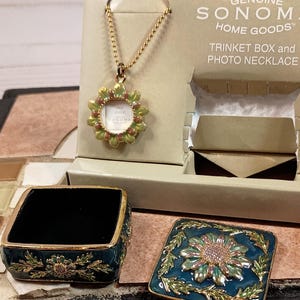 May include: A teal blue trinket box with a gold trim and floral design. The lid is open and shows the interior of the box. The box is part of a set that includes a gold chain necklace with a small picture frame pendant. The box and necklace are packaged in a white box with the text "GENUINE SONOMA HOME GOODS" and "TRINKET BOX and PHOTO NECKLACE".