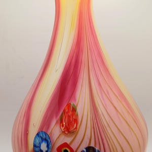 Könnte beinhalten: Eine rosa und gelbe Glasvase mit einer breiten Öffnung und einem schmalen Hals. Die Vase hat ein Spiralmuster und ist mit bunten Glasperlen verziert.