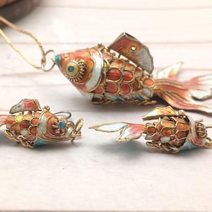 Puede incluir: Un conjunto de joyas de estilo vintage con un colgante y dos pendientes, todos con forma de peces coloridos. Los peces tienen detalles de esmalte naranja, rojo, blanco y azul, con detalles metálicos dorados. El colgante cuelga de una cadena dorada.