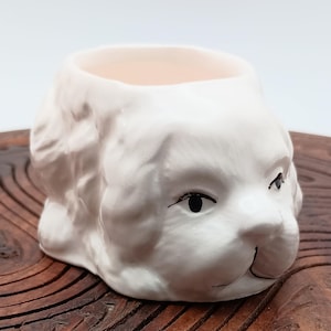 Puede incluir: Huevera de cerámica blanca con forma de cabeza de perro. La taza tiene ojos negros y un diseño simple pintado a mano.