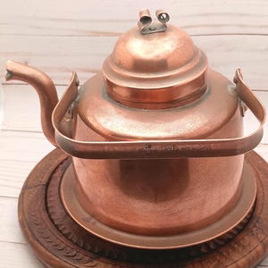 May include: A copper-colored kettle with a rounded body, spout, and lid, sitting on a wooden trivet. The kettle has a handle and a small metal piece on the lid. The text "AVULTUNA 1 1/2 L MADE IN SWEDEN" is visible on the handle.