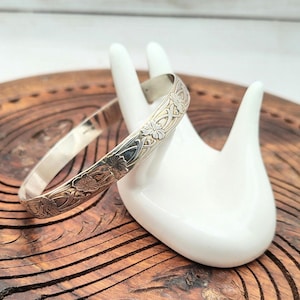 May include: A silver bangle bracelet with an engraved floral design. The bracelet is displayed on a white hand-shaped ceramic holder, set against a wooden background. The floral pattern is detailed and intricate, adding a touch of elegance to the jewelry.