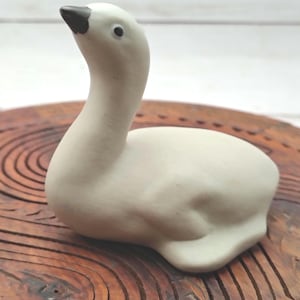 May include: A small, matte white ceramic figurine of a goose in a resting position. The goose has a smooth, rounded body, a long neck curved upwards, and a dark brown beak. A single black dot represents the eye on the side of the head.