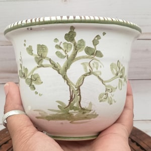 May include: White ceramic planter with a hand-painted tree design in shades of green and brown. The rim features a green and white striped pattern. A decorative piece for indoor or outdoor plants.