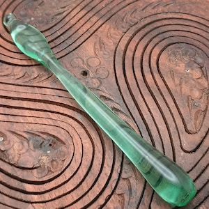 May include: A translucent green glass tool with a rounded tip and a long, slender handle. The tool is approximately 8 inches long. The background is a carved wooden surface with a dark brown finish.