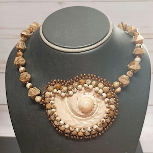 Puede incluir: Un collar artesanal con un gran colgante de concha rosa pálido. El colgante está rodeado de pequeñas cuentas redondas e irregulares en tonos marrón, beige y blanco. El collar tiene una cadena de cuentas con cuentas de tonos similares.