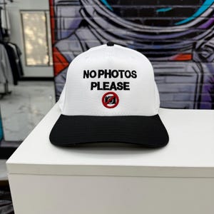 Puede incluir: Gorra de béisbol blanca con visera negra. La gorra tiene el texto "NO PHOTOS PLEASE" en negro, con un círculo rojo y un icono de cámara tachada. La gorra está sobre una superficie blanca.