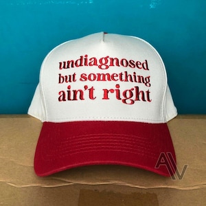 May include: A white baseball cap with a red brim and red embroidered text on the front that reads, "undiagnosed but something ain't right." The cap features a structured front panel and a classic design with a curved visor and stitched detailing.