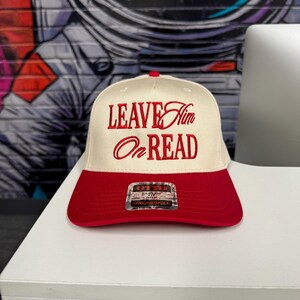 Puede incluir: Una gorra de béisbol blanca con visera roja. La parte delantera de la gorra tiene un texto bordado en rojo que dice "LEAVE Him On READ".
