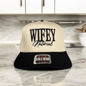 May include: A beige baseball cap with a black brim. The front of the cap has black embroidery that reads "WIFEY Material".