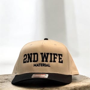 Puede incluir: Gorra de béisbol beige con visera negra. La gorra presenta el texto "2ND WIFE" en letras negras y en negrita, con "MATERIAL" impreso debajo. El sombrero se muestra sobre una superficie clara y texturizada.