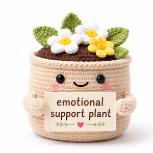 Peut inclure: Un pot de plante beige crocheté avec un visage souriant, tenant une pancarte avec l'inscription "emotional support plant". Le pot est rempli de terre brune et surmonté de fleurs crochetées blanches et jaunes et de feuilles vertes.