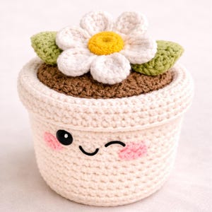Peut inclure: Un pot de fleur crocheté de couleur crème avec un visage souriant. Il présente un dessus marron, des feuilles vertes et une fleur blanche avec un centre jaune. Le pot a un œil qui cligne de l'œil et des joues roses.
