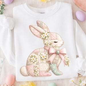 Peut inclure: Sweat-shirt blanc avec un motif de lapin en patchwork. Le lapin est orné de fleurs, d'un nœud rose et d'une pierre précieuse. Le motif utilise des couleurs pastel rose, vert et crème, créant une esthétique douce sur le thème du printemps.