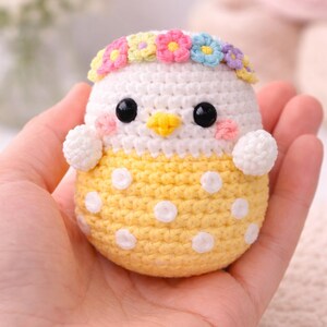 Peut inclure: Un poussin crocheté à la tête blanche et au corps jaune à pois. Il porte une couronne de fleurs, a des yeux noirs et un bec jaune. Le poussin est tenu dans une main. Le travail artisanal est détaillé et coloré.