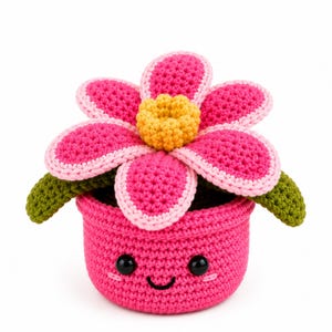 Peut inclure: Une fleur au crochet faite à la main dans un pot. La fleur a des pétales roses avec des bords rose clair et un centre jaune. Le pot rose a un visage souriant et des yeux noirs. Des feuilles vertes sont visibles.