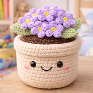 May include: A crocheted flower pot with a smiling face, holding a cluster of purple flowers with yellow centers and green leaves. The pot is beige with a brown top, and the flowers are arranged to look like a bouquet.