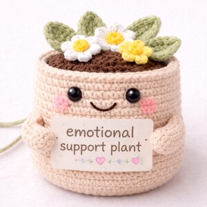Peut inclure: Un pot de plante crocheté avec un visage souriant, des yeux noirs et des joues roses. Le pot contient de la fausse terre, des fleurs blanches et jaunes et des feuilles vertes. Il porte une pancarte avec l'inscription "emotional support plant".