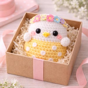 Peut inclure: Un poussin crocheté à la main, orné d'une couronne de fleurs, avec des yeux noirs et un bec jaune, présenté dans une boîte cadeau marron avec du rembourrage en papier. Le corps du poussin est blanc et jaune avec des pois blancs. Un ruban rose entoure la boîte.