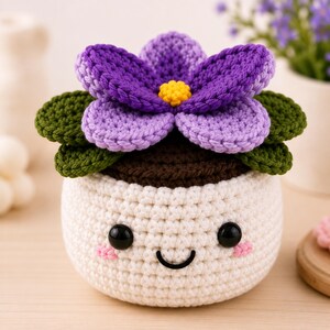 Peut inclure: Un pot de fleur crocheté avec un visage souriant. Le pot est blanc avec un dessus marron, des feuilles vertes et une fleur violette avec un centre jaune. Des yeux en boutons noirs et des joues roses complètent le design.
