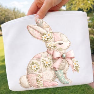Peut inclure: Pochette blanche zippée avec un motif de lapin patchwork. Le lapin est orné d'éléments floraux, d'un nœud rose et d'une gemme turquoise. Le design utilise des couleurs pastel rose, vert et crème, créant une esthétique douce et printanière.