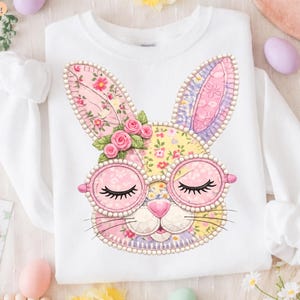 Peut inclure: Sweat-shirt blanc avec un motif de visage de lapin coloré. Le lapin porte des lunettes roses, les yeux fermés et un nez rose. Le visage du lapin est fait de tissu patchwork avec des motifs floraux et des bordures perlées. Une grappe de roses roses orne la tête du lapin.