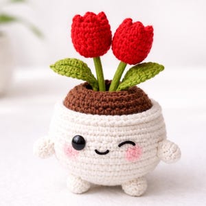 Peut inclure: Un pot crocheté blanc avec un visage souriant, tenant deux tulipes rouges avec des feuilles vertes. Le pot a de petits bras et pieds, et des joues roses. La terre du pot est marron.