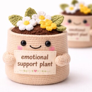 Peut inclure: Pot de plante beige crocheté avec un visage souriant, tenant une pancarte avec l'inscription "emotional support plant". Le pot a des yeux en boutons noirs, des joues roses et un dessus marron avec des fleurs blanches et jaunes crochetées et des feuilles vertes.
