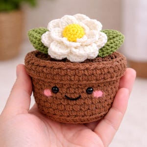 Peut inclure: Un pot de fleur marron crocheté avec un visage souriant, tenant une fleur blanche avec un centre jaune et des feuilles vertes. Le pot mesure environ 7,6 cm de haut.