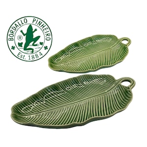 Op de afbeelding: Twee groene keramische serveerschalen in de vorm van bananenbladeren met een getextureerd oppervlak en een kleine lusvormige handgreep aan één uiteinde. Het ronde logo van Bordallo Pinheiro Est. 1884 met een kikker is linksboven zichtbaar.