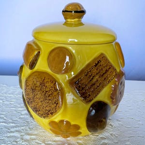 May include: A bright yellow ceramic cookie jar with a matching lid featuring a knob handle. The exterior is decorated with raised, three-dimensional relief designs of various cookies and biscuits in shades of brown, tan, and dark chocolate.